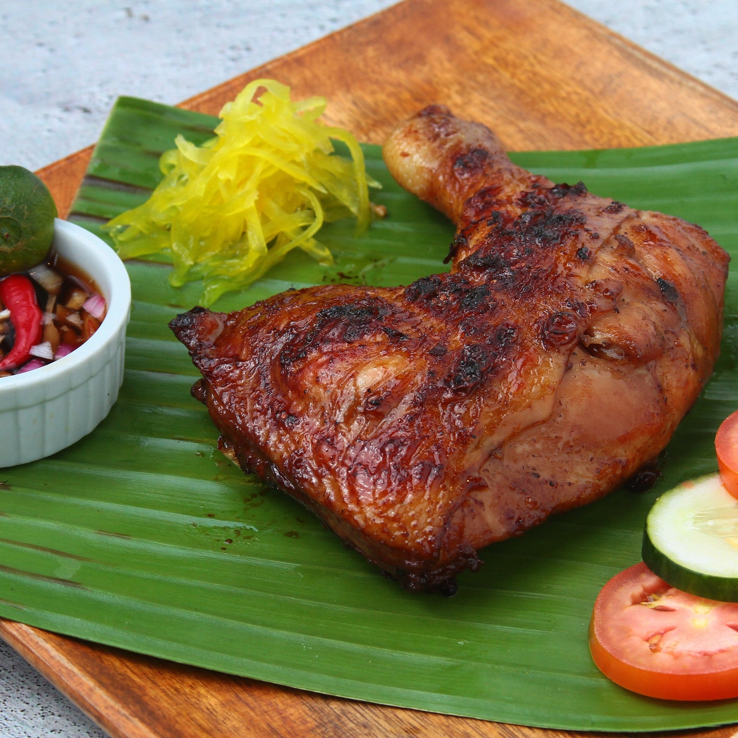 A roasted chicken inasal leg on a banana leaf, accompanied by sliced tomatoes, a lemon wedge, and a small dish of condiment on the side, presented on a wooden board.